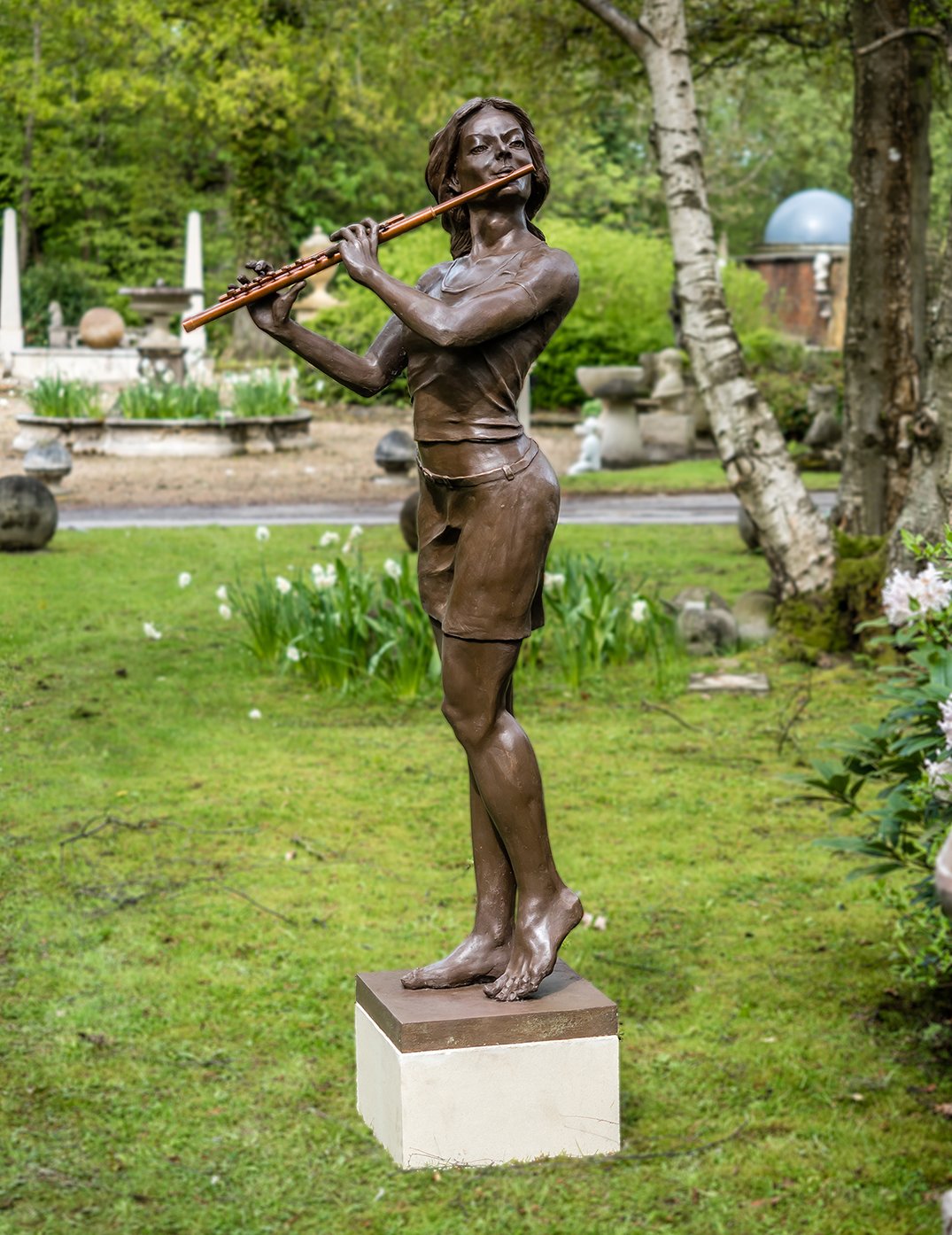 Replica bronze sculpture of a woman playing a flute standing on a concrete base with daffodils in background