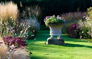 A beautiful garden designed by Kate Ball, featuring swaying grasses and perennials around a Chilstone traditional stone garden planter with basket weave design and lions feet, raised on a plinth as the central feature.