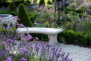 A curved stone bench, handmade by Chilstone, engraved as a feature in a landscaped garden design, with richly planted beds with purple verbena and lavender. 